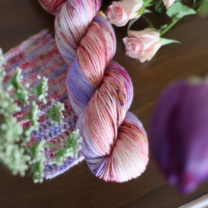 DK yarn and crochet and knit swatches colored different shades of light and dark pink with creme colors and a light lavender. This is on top of a brown table with some purple and light pink tulips around the yarn and babies breath flowers too.