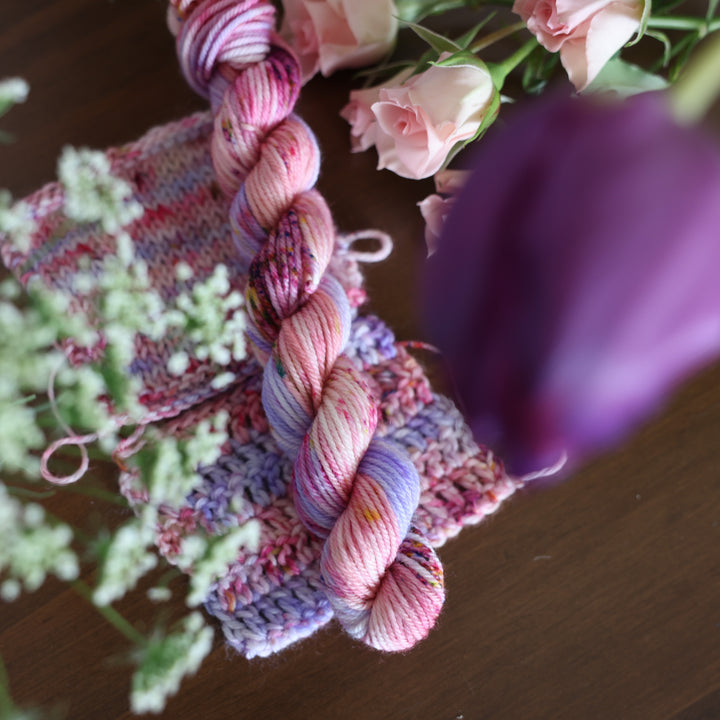 DK mini yarn and crochet and knit swatches colored different shades of light and dark pink with creme colors and a light lavender. This is on top of a brown table with some purple and light pink tulips around the yarn.