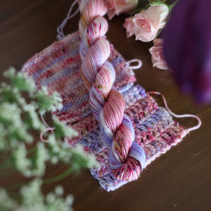 Fingering mini yarn and crochet and knit swatches colored different shades of light and dark pink with creme colors and a light lavender. This is on top of a brown table with some purple and light pink tulips around the yarn.