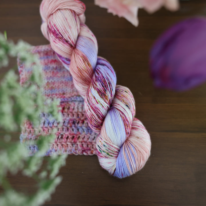 Silk yarn and crochet and knit swatches colored different shades of light and dark pink with creme colors and a light lavender. This is on top of a brown table with some purple and light pink tulips around the yarn.