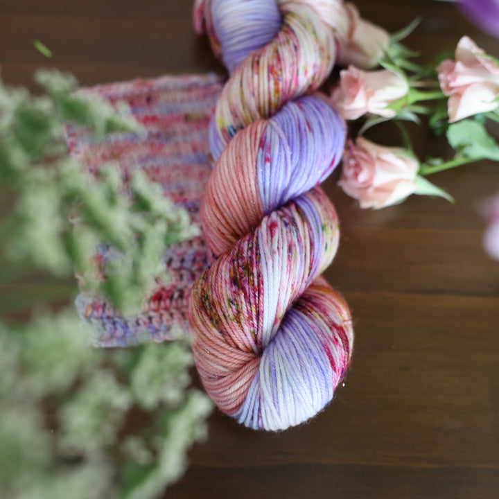 Sparkle DK yarn and crochet and knit swatches colored different shades of light and dark pink with creme colors and a light lavender. This is on top of a brown table with some purple and light pink tulips around the yarn and some babies breath flowers too.