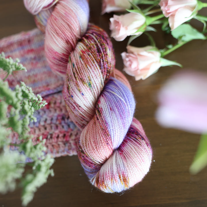Fingering yarn and crochet and knit swatches colored different shades of light and dark pink with creme colors and a light lavender. This is on top of a brown table with some purple and light pink tulips around the yarn.