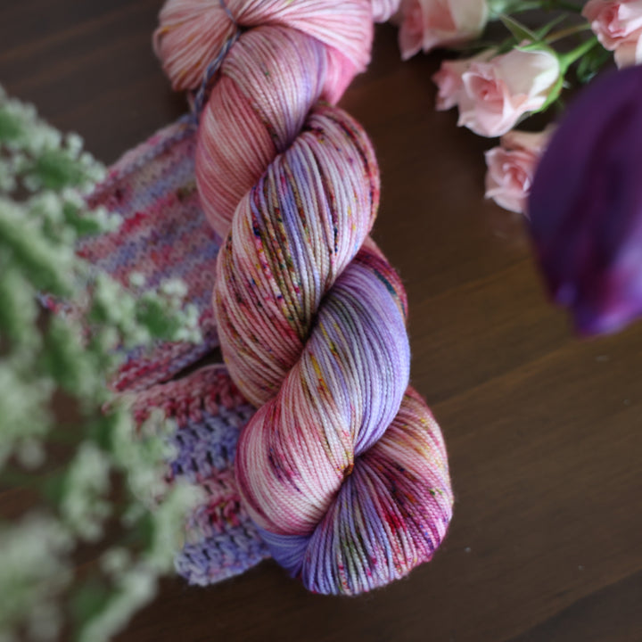 Sport yarn and crochet and knit swatches colored different shades of light and dark pink with creme colors and a light lavender. This is on top of a brown table with some purple and light pink tulips around the yarn.