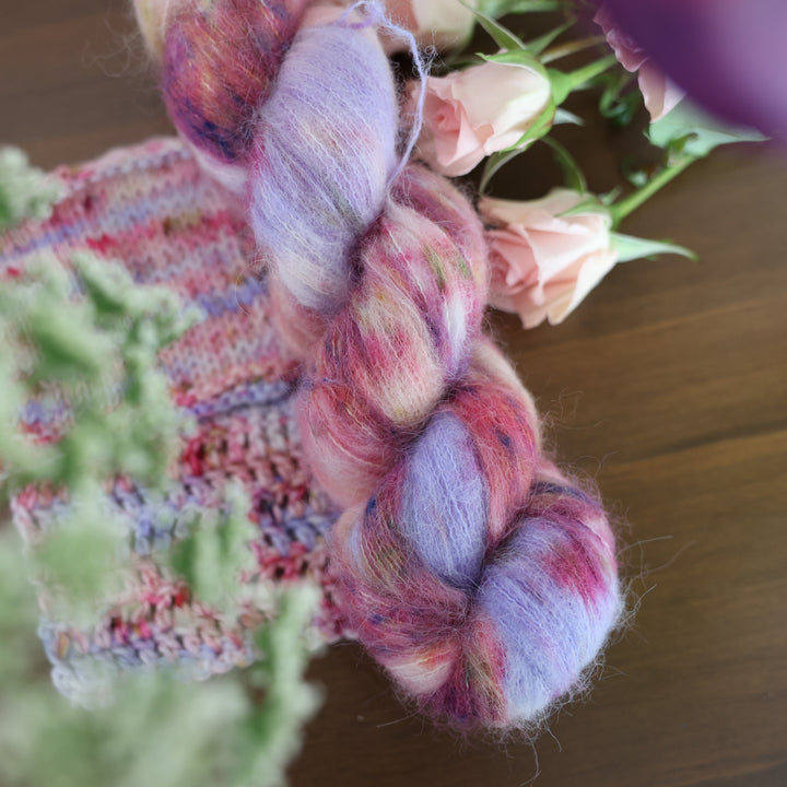 Suri yarn and crochet and knit swatches colored different shades of light and dark pink with creme colors and a light lavender. This is on top of a brown table with some purple and light pink tulips around the yarn.