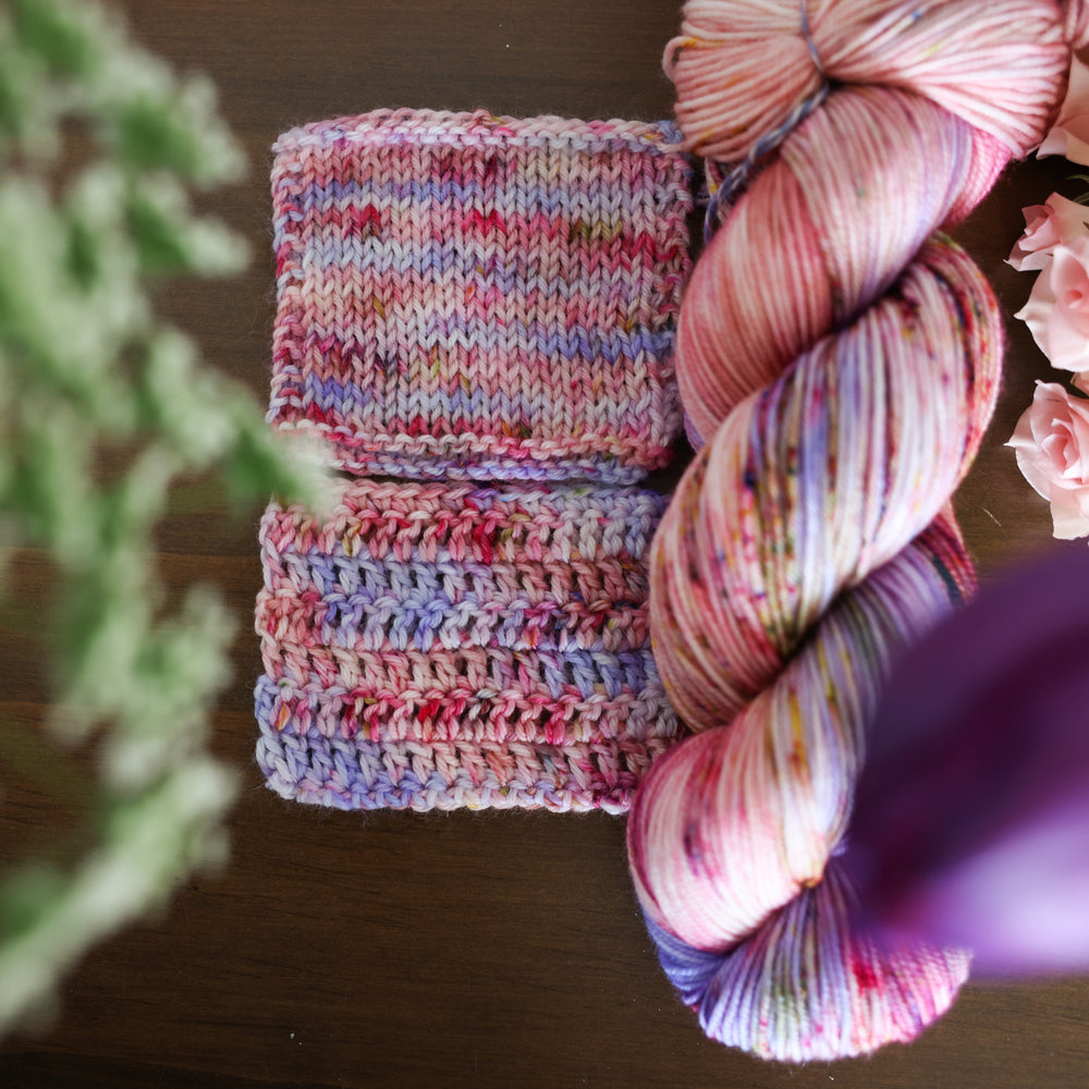 Yarn skein and crochet and knit swatches colored different shades of light and dark pink with creme colors and a light lavender. This is on top of a brown table with some purple and light pink tulips around the yarn.