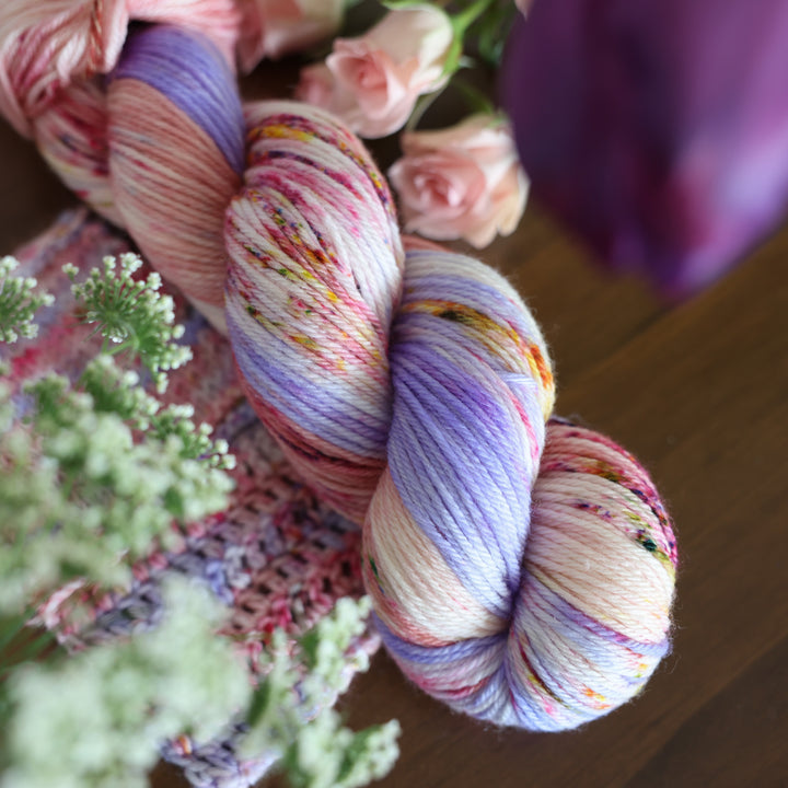 Worsted yarn and crochet and knit swatches colored different shades of light and dark pink with creme colors and a light lavender. This is on top of a brown table with some purple and light pink tulips around the yarn.