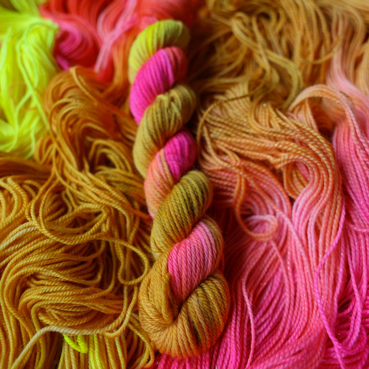 DK mini yarn colored a bright yellow, pink, and a salmon pink orange, also a yellow beige color. This skein is on top of unwound skeins of this colorway.