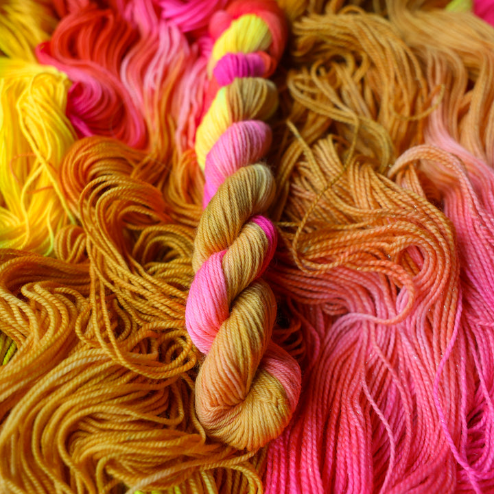 Fingering mini yarn colored a bright yellow, pink, and a salmon pink orange, also a yellow beige color. This skein is on top of unwound skeins of this colorway.