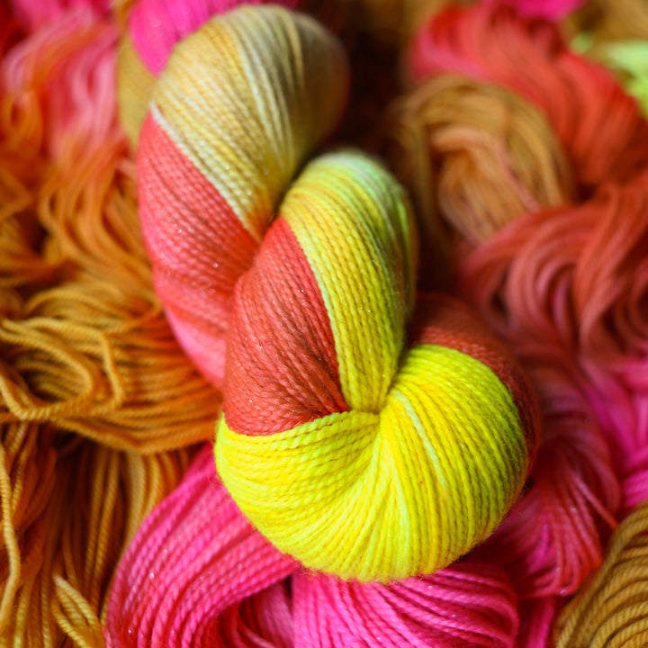 Sparkle fingering yarn colored a bright yellow, pink, and a salmon pink orange, also a yellow beige color. This skein is on top of unwound skeins of this colorway.