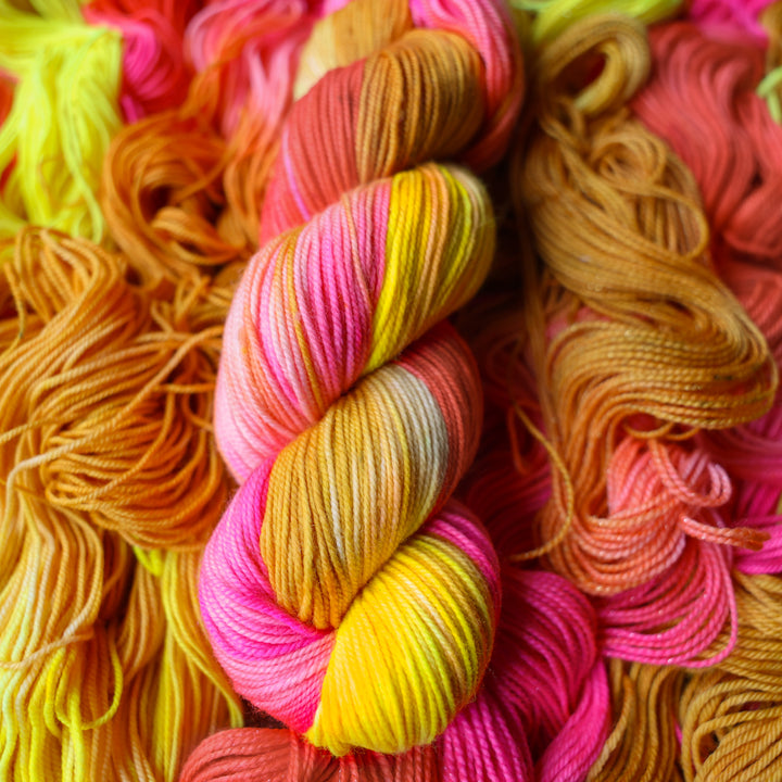 Sport yarn colored a bright yellow, pink, and a salmon pink orange, also a yellow beige color. This skein is on top of unwound skeins of this colorway.