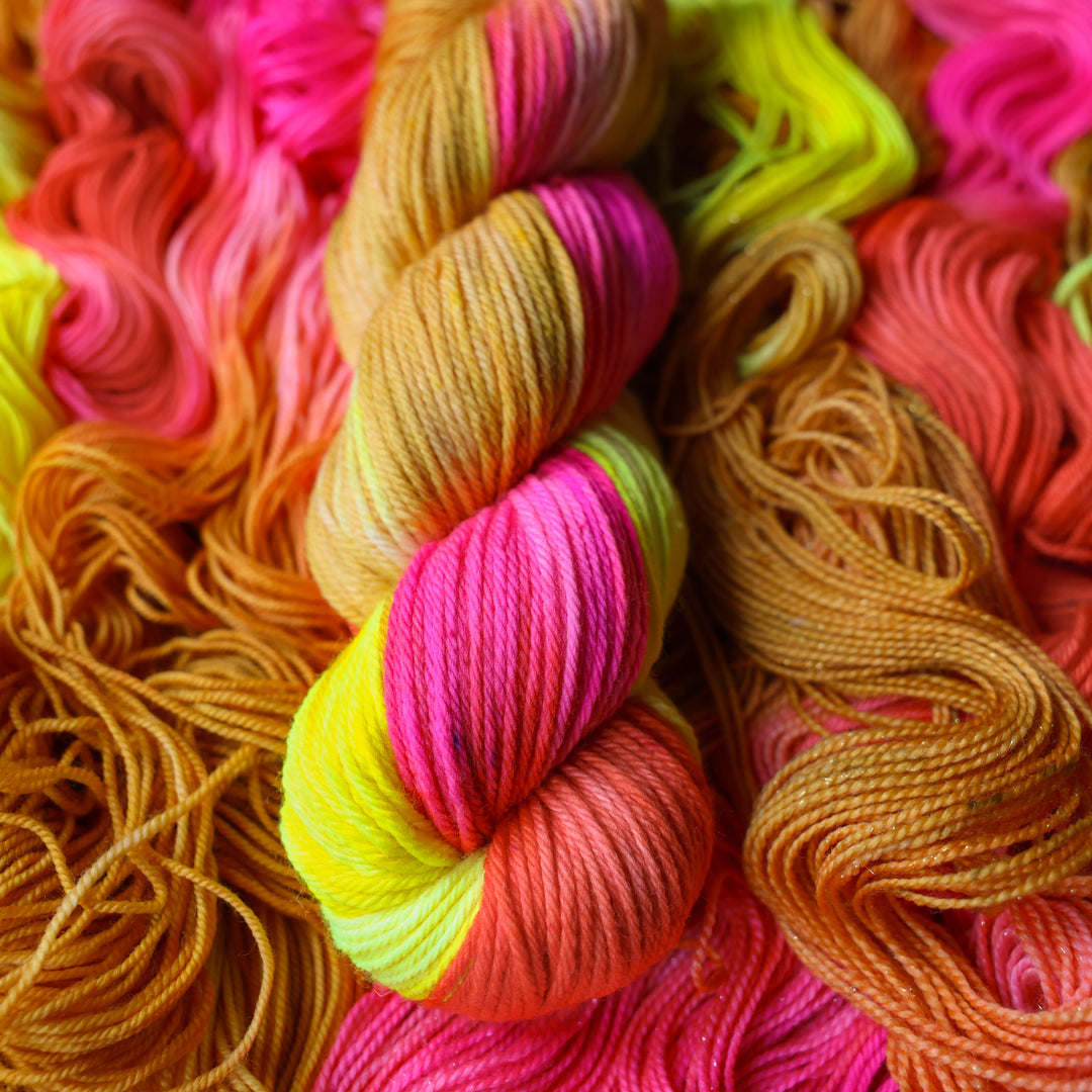 Worsted yarn colored a bright yellow, pink, and a salmon pink orange, also a yellow beige color. This skein is on top of unwound skeins of this colorway.