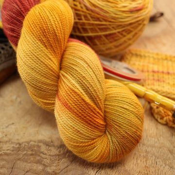 A yellow and orange/pink fingering yarn on top of a wooden table and a caked version of this yarn with and a pen, ribbon, and swatches of that yarn on the side.