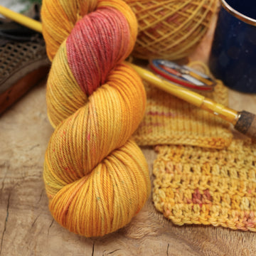 A yellow and orange/pink Dk yarn on top of a wooden table and a caked version of this yarn with and a pen, ribbon, and a mug next to it.