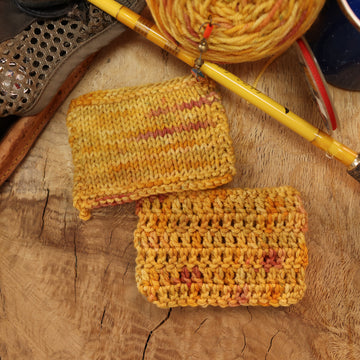 A yellow and orange/pink yarn in crochet and knit swatches on top of a wooden table and a caked version of this yarn with and a pen, ribbon, and a mug next to it.