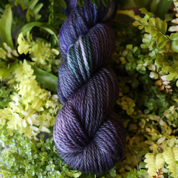 Bulky yarn colored dark purple and some dark blue. This skein is on top of a bed of ferns and plants