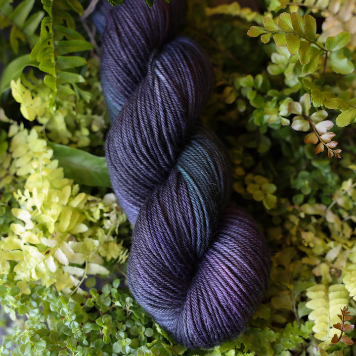 DK yarn colored dark purple and some dark blue. This skein is on top of a bed of ferns and plants