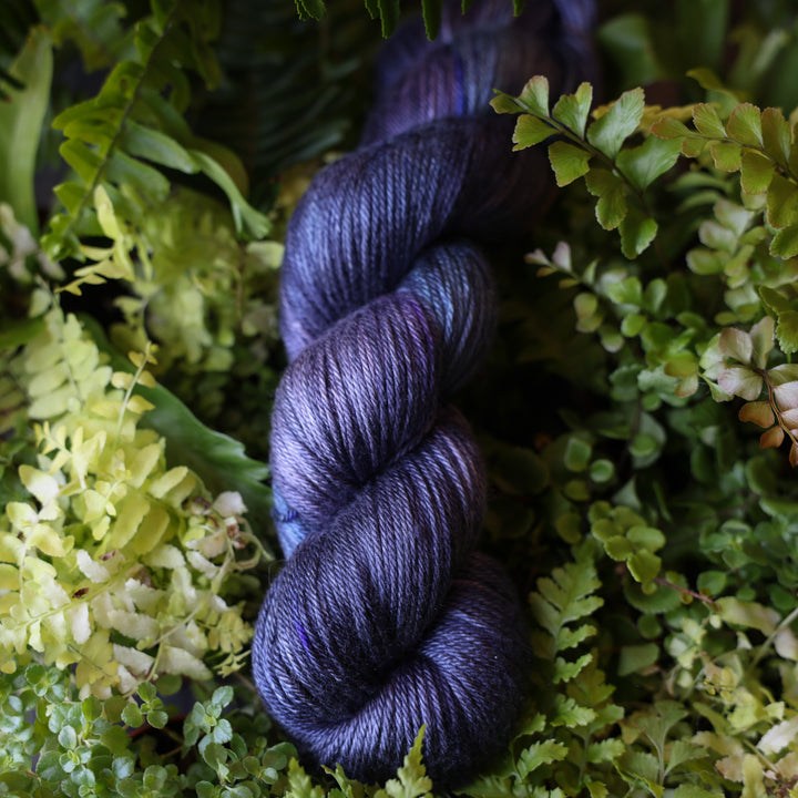 DK silk yarn colored dark purple and some dark blue. This skein is on top of a bed of ferns and plants