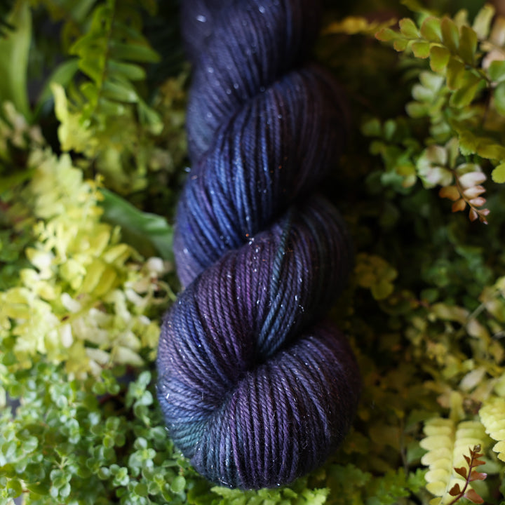Sparkle DK yarn colored dark purple and some dark blue. This skein is on top of a bed of ferns and plants