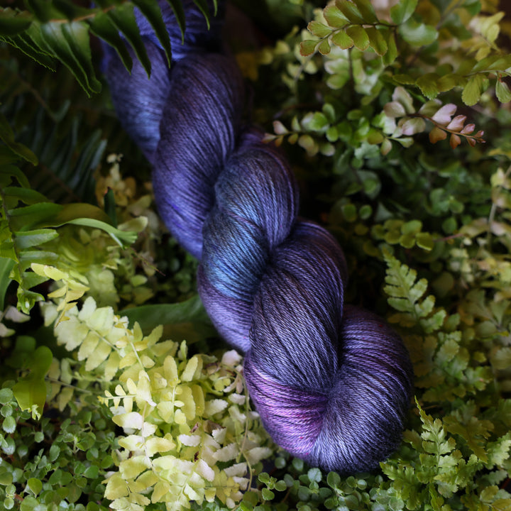 Silk yarn colored dark purple and some dark blue. This skein is on top of a bed of ferns and plants