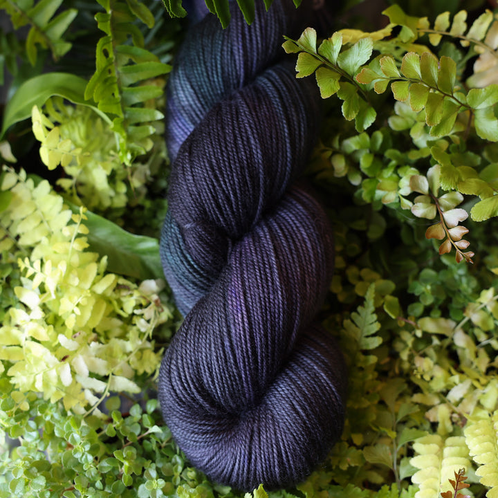 Sport yarn colored dark purple and some dark blue. This skein is on top of a bed of ferns and plants