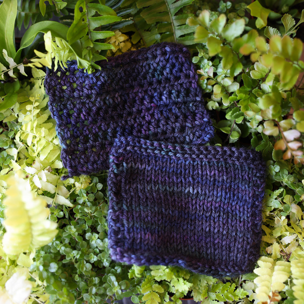 Crochet and knit swatches colored dark purple and some dark blue. These swatches are on top of a bed of ferns and plants