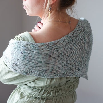 Women turned away wearing a Twisted Whispers Knit Shawl. The Shawl is light blue with ints of darker blue and lavender.