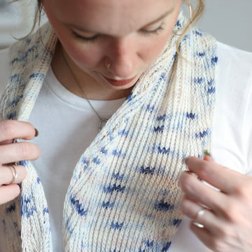 Women looking down, wearing a Twisted Whispers Knit Shawl. The Shawl is white with hints of light blue details.