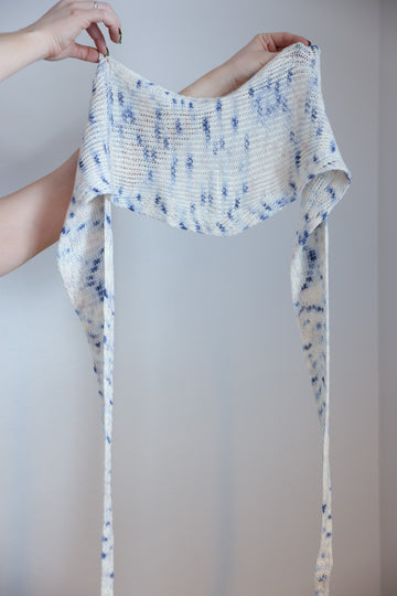 A person holding  a Twisted Whispers Knit Shawl. The Shawl is white with hints of light blue details.