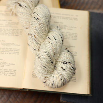 White with black and brown speckles. Fingering weight on top of an opened book with other books almost fully in the picture, all on top of a dark wooden table.