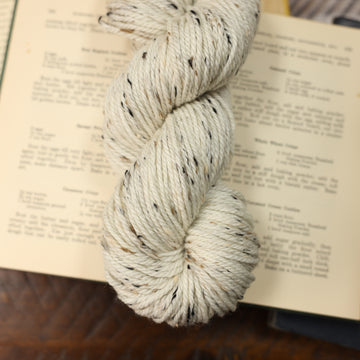 White and black and brown speckled square yarn on top of an opened book with other books almost fully in the picture, all on top of a dark wooden table.