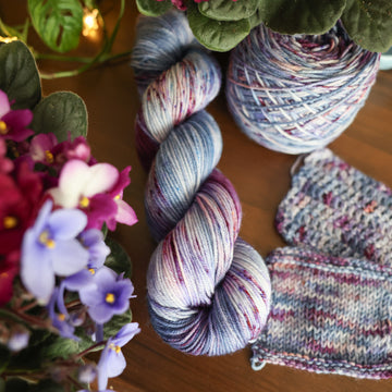One DK skein of yarn colored a lovely light lavender with delicate white blush and dark purple speckling, Violet is delicate with a victorian floral mood. All sitting on top of a wooden table surrounded by colorful flowers. Off to the side there is one knit and one crochet swatches and a caked skein.
