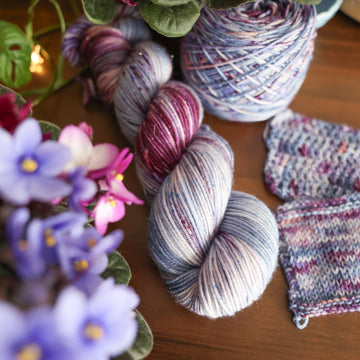 One DK Sport skein of yarn colored a lovely light lavender with delicate white blush and dark purple speckling, Violet is delicate with a victorian floral mood. All sitting on top of a wooden table surrounded by colorful flowers. Off to the side there is one knit and one crochet swatches and a caked skein.
