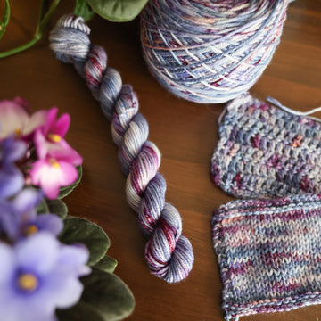 One DK Mini skein of yarn colored a lovely light lavender with delicate white blush and dark purple speckling, Violet is delicate with a victorian floral mood. All sitting on top of a wooden table surrounded by colorful flowers. Off to the side there is one knit and one crochet swatches and a caked skein.
