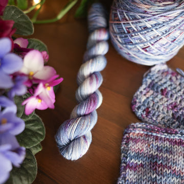 One Fingering Mini skein of yarn colored a lovely light lavender with delicate white blush and dark purple speckling, Violet is delicate with a victorian floral mood. All sitting on top of a wooden table surrounded by colorful flowers. Off to the side there is one knit and one crochet swatches and a caked skein.
