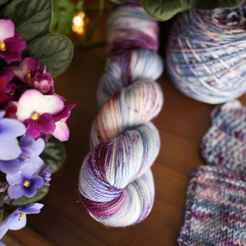 One Sparkle fingering skein of yarn colored a lovely light lavender with delicate white blush and dark purple speckling, Violet is delicate with a victorian floral mood. All sitting on top of a wooden table surrounded by colorful flowers. Off to the side there is one knit and one crochet swatches and a caked skein.
