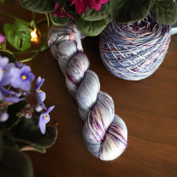 One Silk skein of yarn colored a lovely light lavender with delicate white blush and dark purple speckling, Violet is delicate with a victorian floral mood. All sitting on top of a wooden table surrounded by colorful flowers. Off to the side there is a caked skein.
