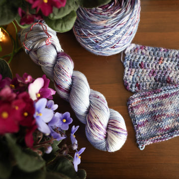 One Silk skein of yarn colored a lovely light lavender with delicate white blush and dark purple speckling, Violet is delicate with a victorian floral mood. All sitting on top of a wooden table surrounded by colorful flowers. Off to the side there is one knit and one crochet swatches and a caked skein.
