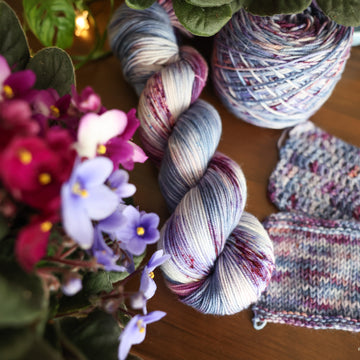 One Sport skein of yarn colored a lovely light lavender with delicate white blush and dark purple speckling, Violet is delicate with a victorian floral mood. All sitting on top of a wooden table surrounded by colorful flowers. Off to the side there is one knit and one crochet swatches and a caked skein.
