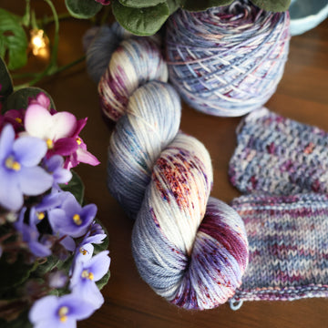 One Worsted skein of yarn colored a lovely light lavender with delicate white blush and dark purple speckling, Violet is delicate with a victorian floral mood. All sitting on top of a wooden table surrounded by colorful flowers. Off to the side there is one knit and one crochet swatches and a caked skein.
