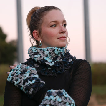 A photo of a women from the front wearing Voidbane Cowl and Mitts, a dragon scale crochet with icy blue, grey, dark blue and black colors.