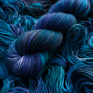 DK yarn colored with different shades of blue and some light bluish purple. Unwound weights of this same yarn is underneath this skein.