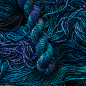 Fingering Mini yarn colored with different shades of blue and some light bluish purple. Unwound weights of this same yarn is underneath this skein.