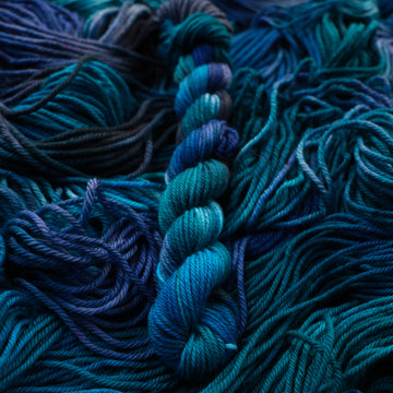 DK mini yarn colored with different shades of blue and some light bluish purple. Unwound weights of this same yarn is underneath this skein.
