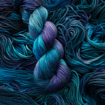 Silk yarn colored with different shades of blue and some light bluish purple. Unwound weights of this same yarn is underneath this skein.
