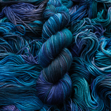 Sport yarn colored with different shades of blue and some light bluish purple. Unwound weights of this same yarn is underneath this skein.