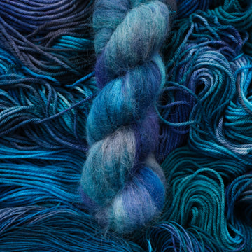 Suri yarn colored with different shades of blue and some light bluish purple. Unwound weights of this same yarn is underneath this skein.