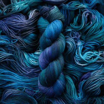 Fingering yarn colored with different shades of blue and some light bluish purple. Unwound weights of this same yarn is underneath this skein.