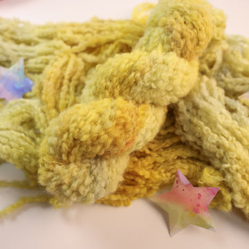 A fluffy yellow yarn skein on top of an unwound version of that yarn, on top of a white table, with a couple of multicolored stars around the yarn.