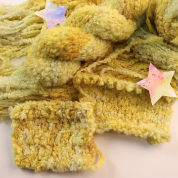A fluffy yellow yarn skein on top of an unwound version of that yarn, on top of a white table, with a couple of multicolored stars around the yarn, and knit and crochet swatches of the yellow yarn next to the other yarn.