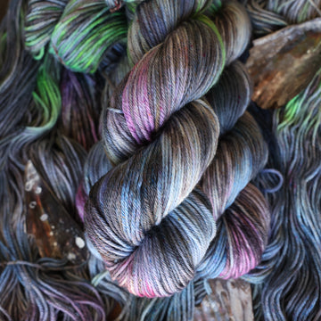 Worsted yarn colored with different shades of grey and streaks of green, and dark pink, also there are also blue and purple speckles. These skeins are on top of unwound skeins of the same color way with some shells on top.